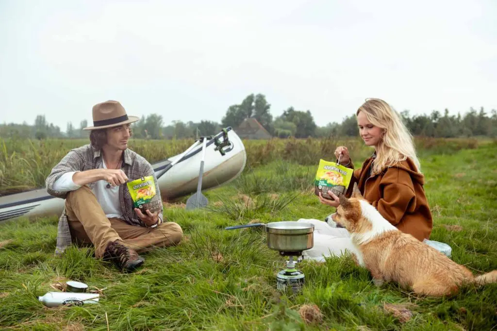 couple et chien en camping, avec un canoë et dégustant un repas d’aventure