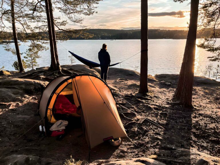 Une tente-dôme éclairée sur un site de bivouac sauvage rocailleux, au bord d’un grand lac en Suède, lors d’un coucher de soleil chaleureux.